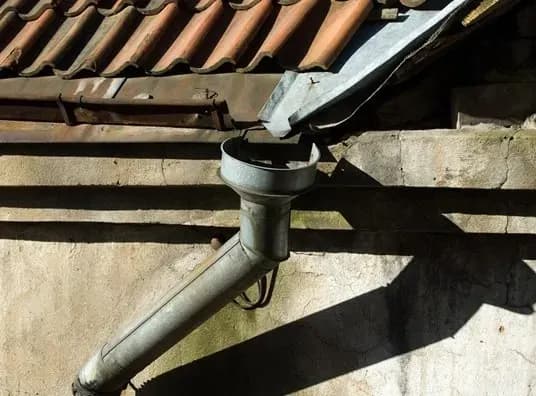 Old roof with weathered tiles and metal gutter pipe casting shadows on a sunlit, aged wall. The scene conveys a rustic and vintage feel.