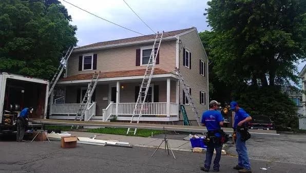 A two-story house with beige siding has ladders propped against it. Workers in blue shirts, tools at hand, are preparing for roof renovations.