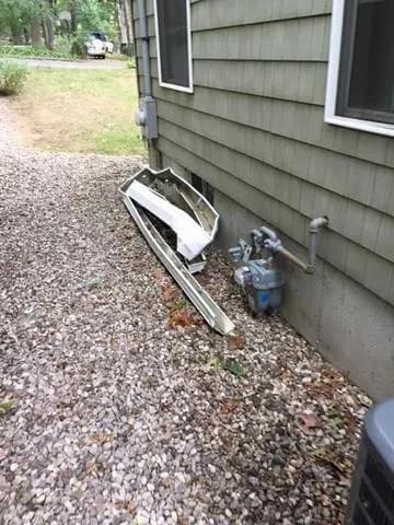 Damaged metal siding leans against a house with gray-green paneling, next to a gas meter on a gravel path. The scene suggests recent damage.