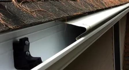 Close-up of a clean, white aluminum gutter under a shingled roof with pine needles. The scene conveys a sense of cleanliness and maintenance.
