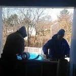 Two workers in winter attire install a window frame. They are silhouetted against a bright outdoors with bare trees and houses in the background.