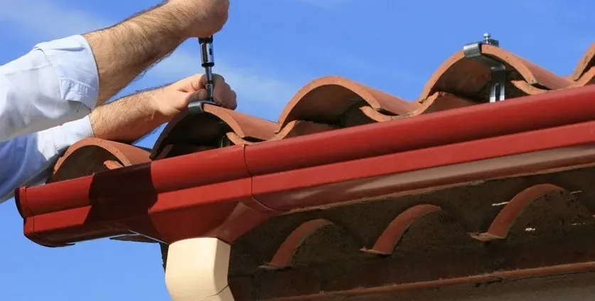 Hands using a screwdriver to adjust metal clamps on curved terracotta roof tiles. The scene conveys careful maintenance under a clear blue sky.