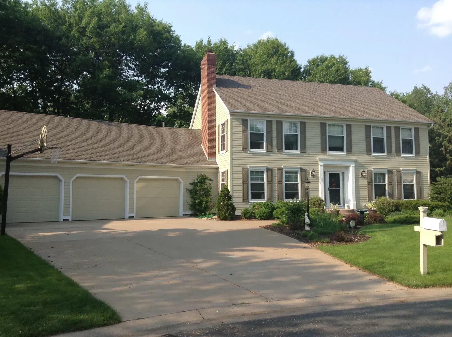 A two-story suburban house with beige siding, white trim, and dark shutters. It features a three-car garage, a manicured lawn, and a basketball hoop.