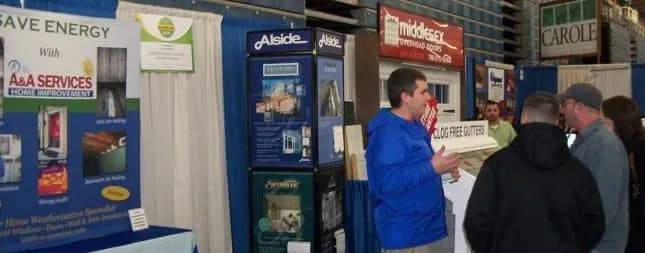 An indoor home improvement expo with booths featuring energy-saving solutions and gutter systems. A presenter engages four attendees, creating an informative and engaged atmosphere.