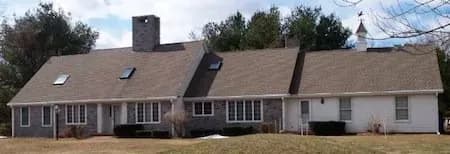 A large, single-story, gray brick house with multiple skylights sits amid trees. It has a steep roof, central chimney, and a weather vane on the right.