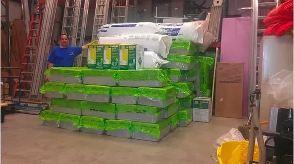 A man stands in a storage area next to a large stack of green-wrapped insulation materials. Ladders and various equipment are visible in the background.