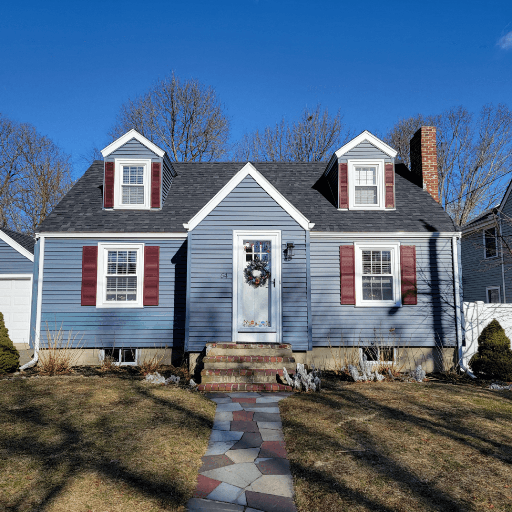 A charming blue house with white trim and a steep gray roof. It features maroon shutters, a front door with a wreath, and a stone walkway. The sky is clear and blue.