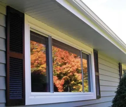 A white-framed window on a house reflects vibrant autumn trees, showcasing shades of orange and red. Dark shutters contrast with the light siding.