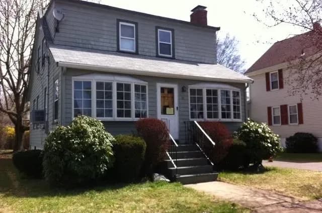 A two-story gray house with large front windows, a small porch, and shrubbery. The scene conveys a suburban, quiet, and inviting atmosphere.