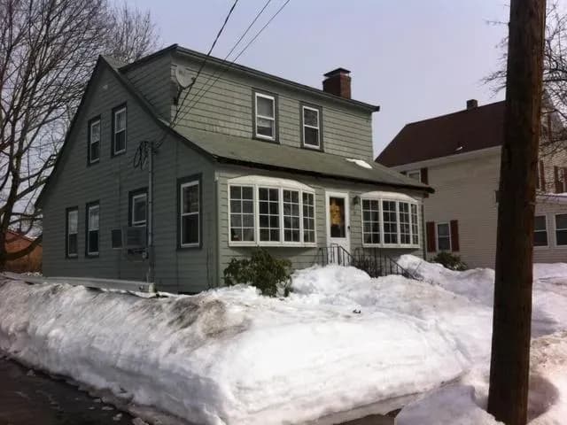 Gray house with snow-covered yard, surrounded by large snowbanks under a cloudy sky. Tree branches and neighboring homes are visible, conveying a chilly winter atmosphere.