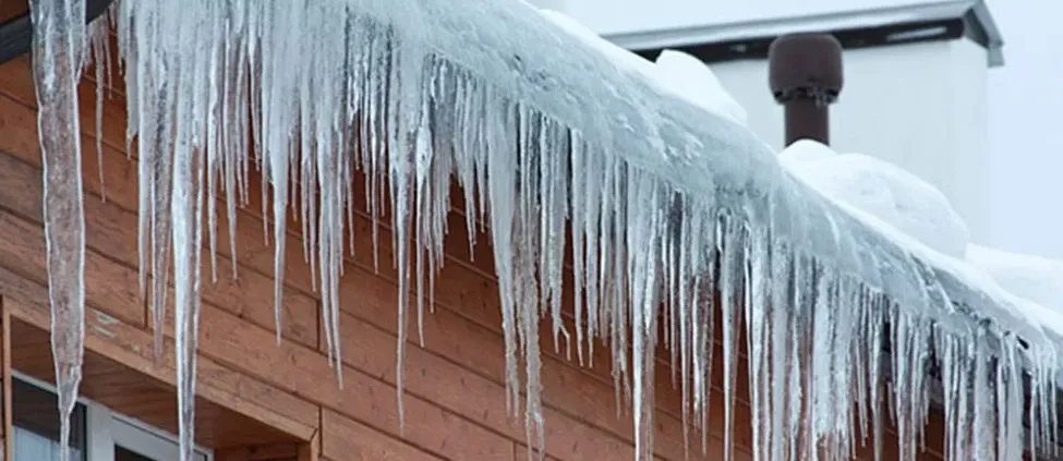 Icicles hang from a snow-covered wooden roof against a cloudy sky. The scene conveys a cold, wintry atmosphere with crisp, clear ice formations.