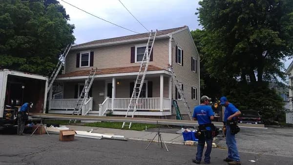 Two workers in blue shirts stand outside a beige two-story house with ladders propped against it. There's construction equipment and a truck nearby.