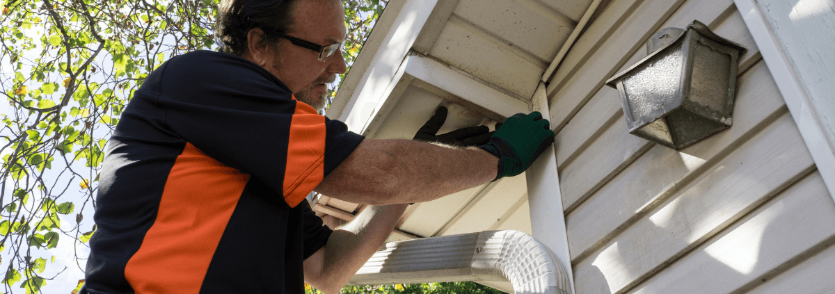 A man in a black and orange shirt and green gloves cleans a house gutter, focused and careful, on a sunny day beside light gray siding and a lantern.