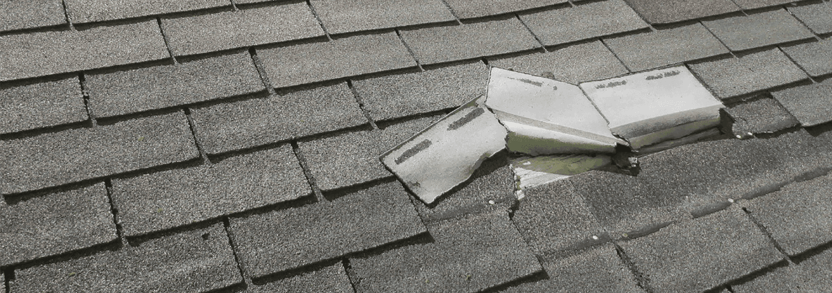 Damaged gray asphalt shingle roof with three shingles upturned, exposing the underlying structure. The image conveys a need for repair.