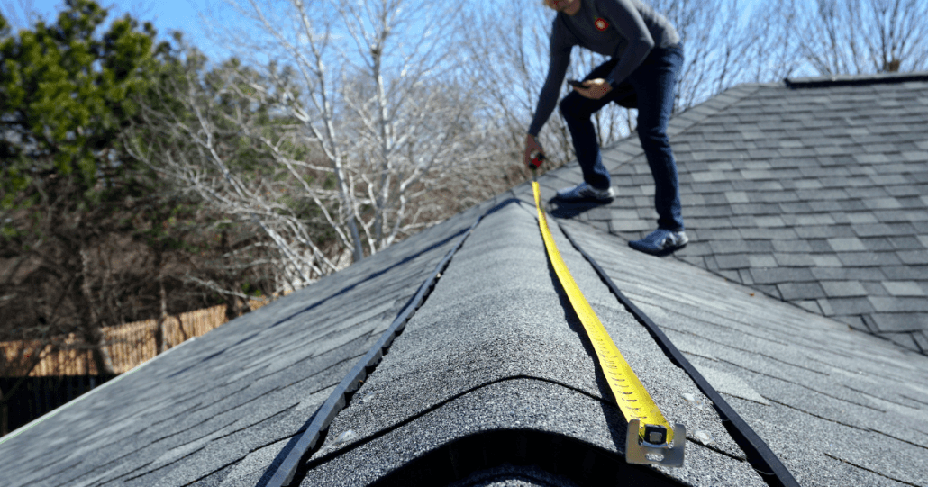 A person measures the peak of a shingled roof with a yellow tape measure. Bare trees and evergreen branches are visible under the clear blue sky.