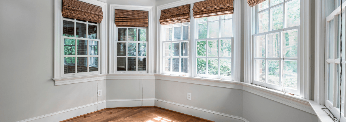 Bright room with bay windows featuring brown woven shades, light gray walls, and a wooden floor. Sunlight streams inside, creating a serene atmosphere.