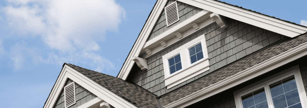 Gabled roof and upper story of a house with greenish-gray siding and white-trimmed windows, set against a blue sky with scattered clouds.