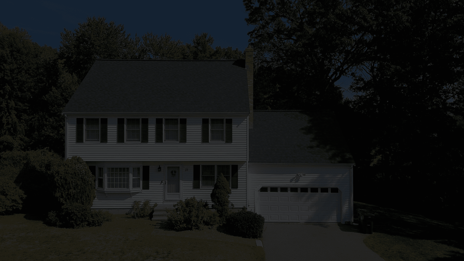 A two-story house with white siding and black shutters is seen. It has a dark gray roof, a covered porch, and a two-car garage, surrounded by trees.