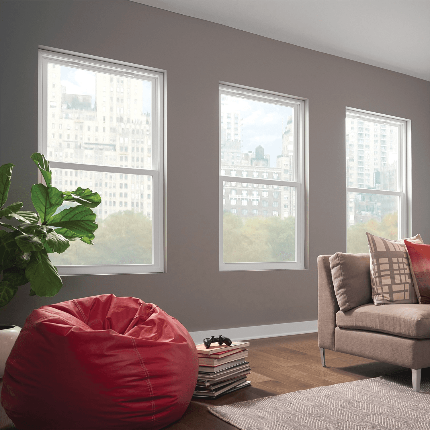 Modern living room with large windows showing a cityscape. Includes a red bean bag, beige sofa, green plant, and stacked books on a wooden floor. Cozy and inviting.