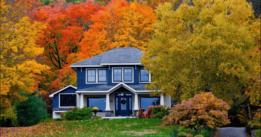 A charming two-story house with a dark blue exterior is surrounded by vibrant autumn foliage in shades of red, orange, and yellow, creating a cozy, serene atmosphere.