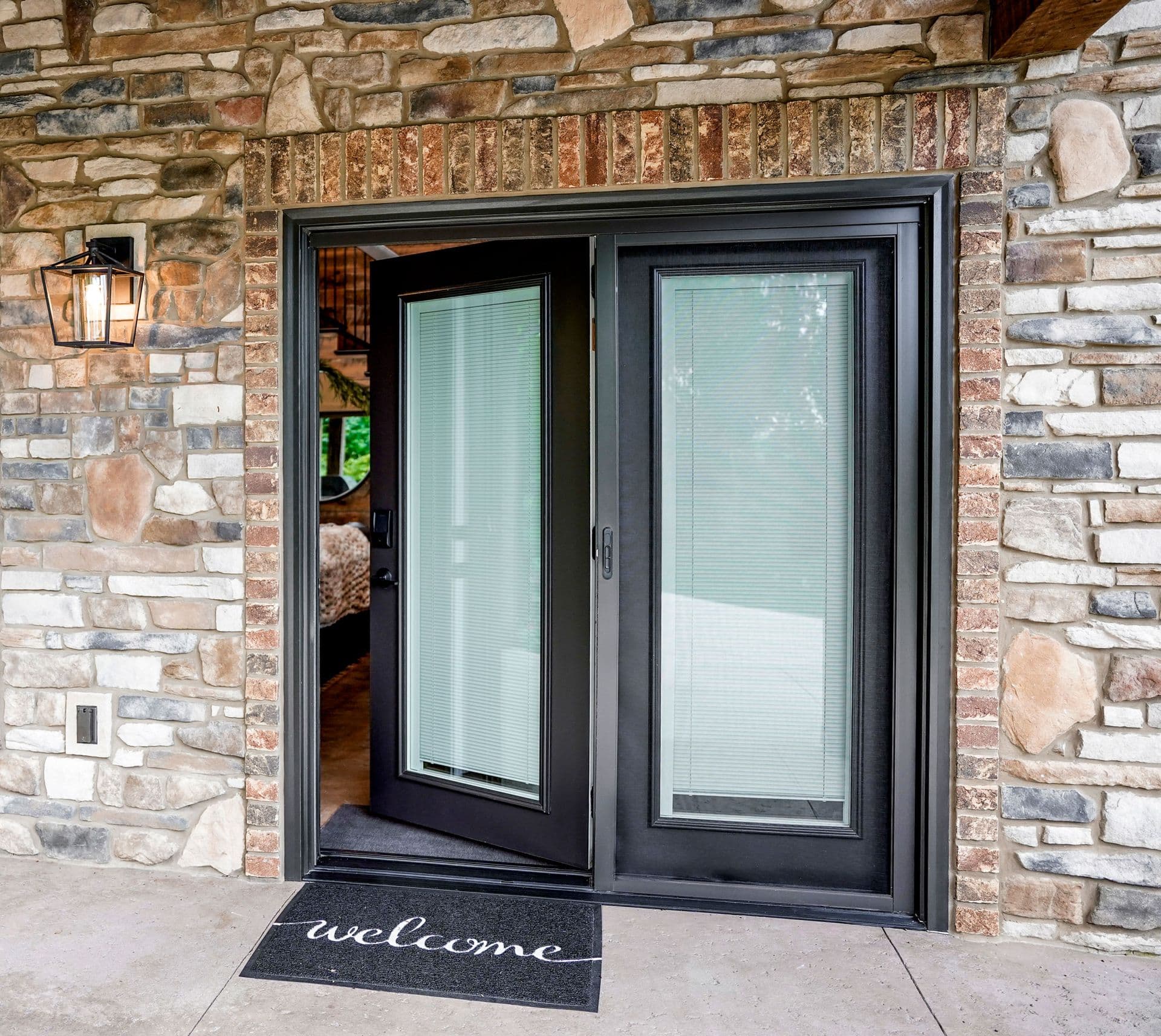 Stone entryway with a partially open black-framed glass door, flanked by a lantern. "Welcome" mat on the ground conveys warmth and hospitality.