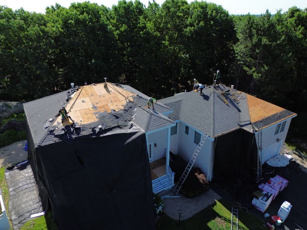 Aerial view of workers repairing a large house roof covered with tarps and shingles, surrounded by ladders, in a lush, tree-lined area.