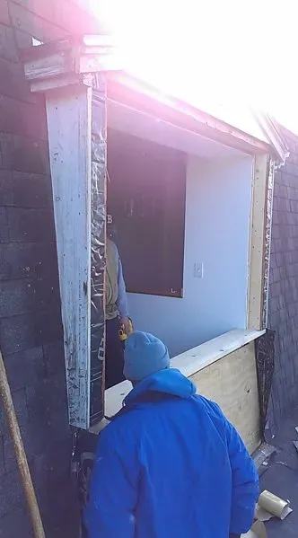 A construction worker in a blue jacket and hat stands outside a building with an open wall, restoring a missing section. Sunlight casts a glare, creating a bright, focused mood.
