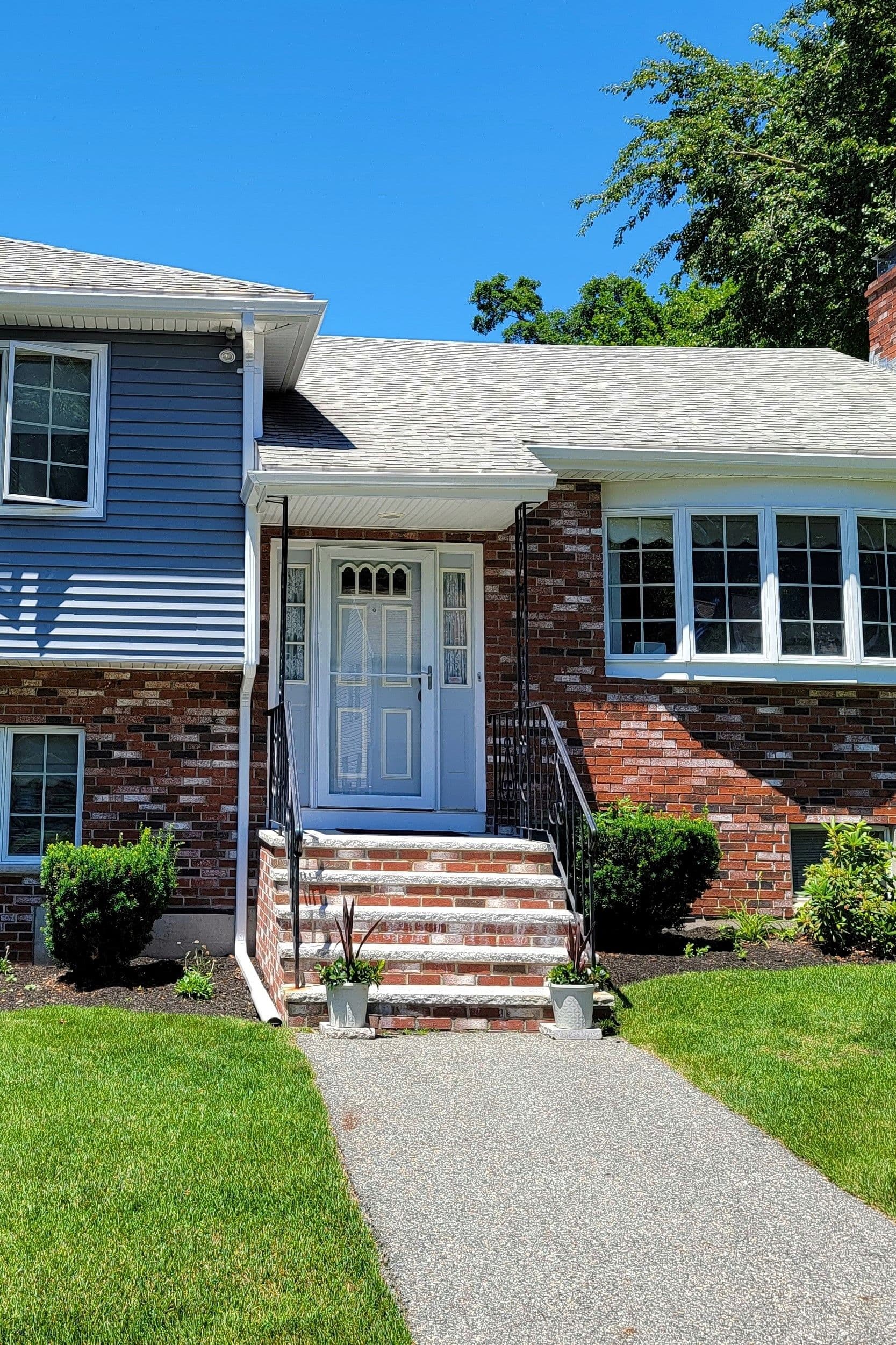 Front view of a suburban house with a brick facade, white door, and steps flanked by potted plants. Manicured lawn and clear, sunny sky enhance the inviting feel.