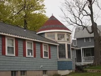 A quaint blue house features a distinctive conical turret with large windows and a red roof. Nearby, a bare tree and another house with a porch are visible.