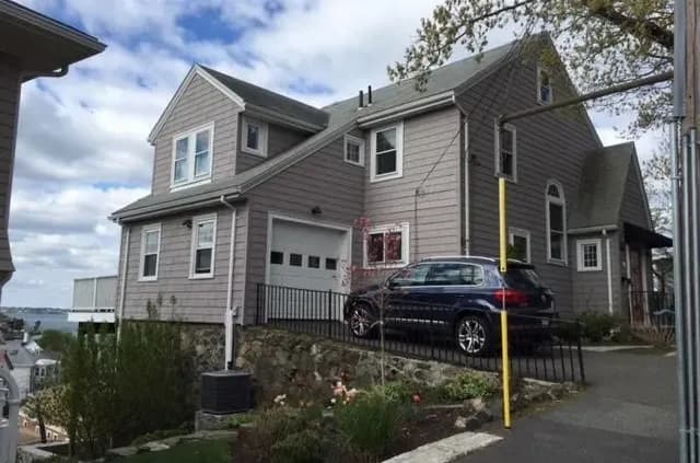 A two-story gray house on a hilltop, featuring a blue car parked on the sloped driveway. A railing surrounds the drive. Ocean view in background.