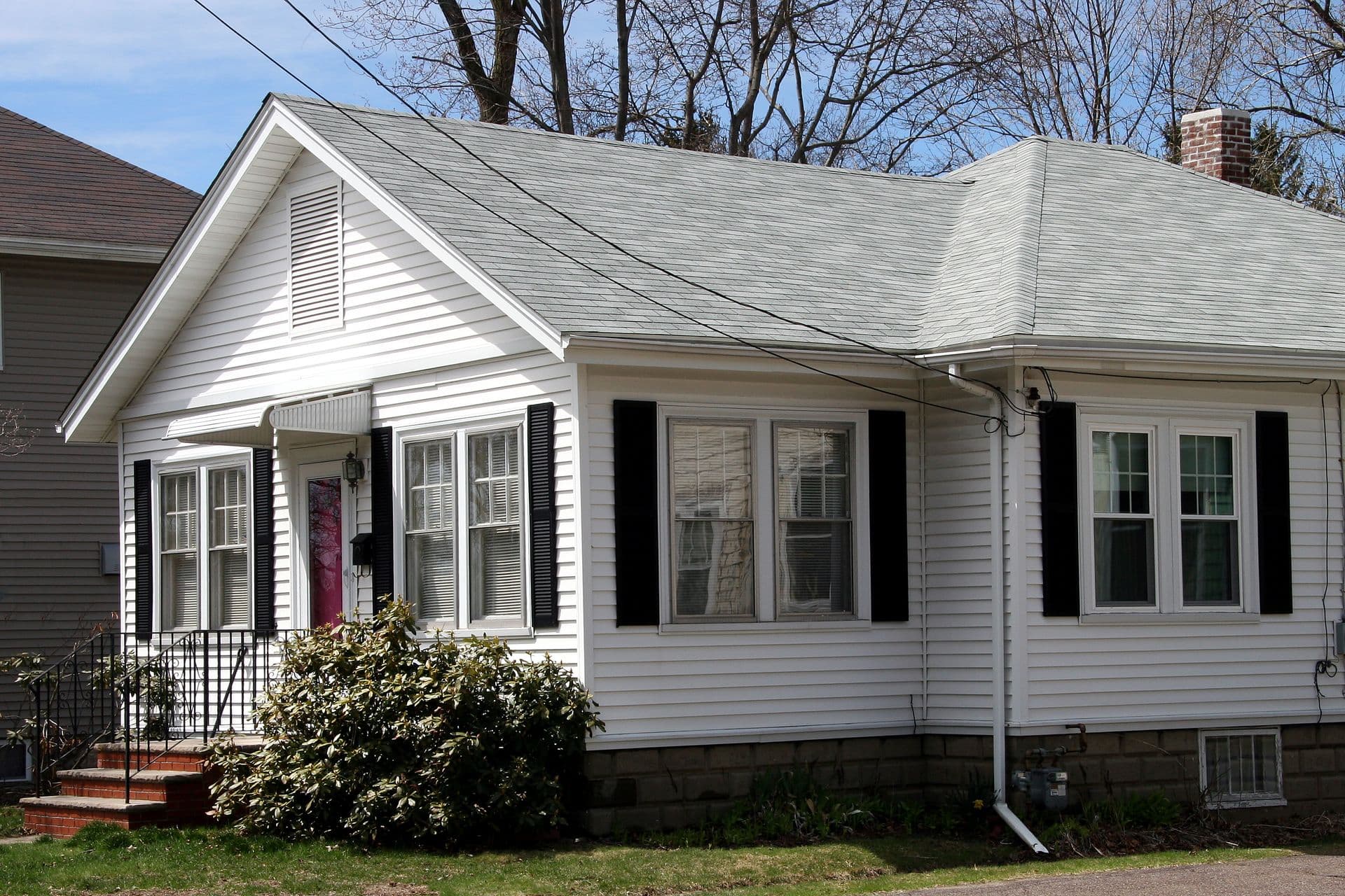 A cozy, single-story white house with a gray gabled roof and black shutters, surrounded by bare trees. A small porch with bushes adds charm.