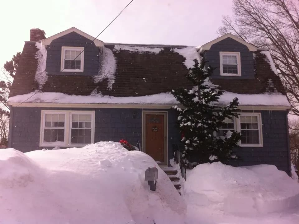 A snow-covered blue house with a steep roof. Large piles of snow surround the entrance and a tree stands to the right, creating a peaceful, wintry scene.
