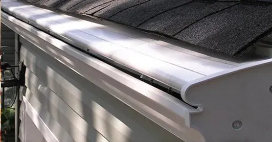 Close-up of a clean, white gutter installed beneath dark roof shingles, casting shadows on light siding. It conveys a sense of neatness and functionality.