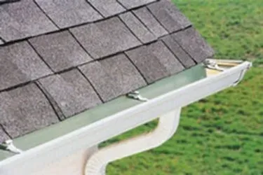 Close-up of a rooftop corner with gray shingles and a white rain gutter. The green grass lawn below adds contrast to the structured roof design.