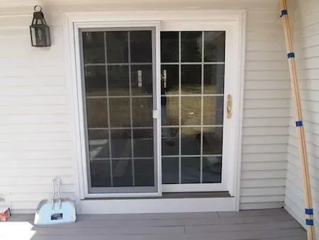 Sliding glass door with grid pattern on a white-paneled exterior wall. A dustpan sits on the left; wooden poles lean on the right. Bright and tidy setting.