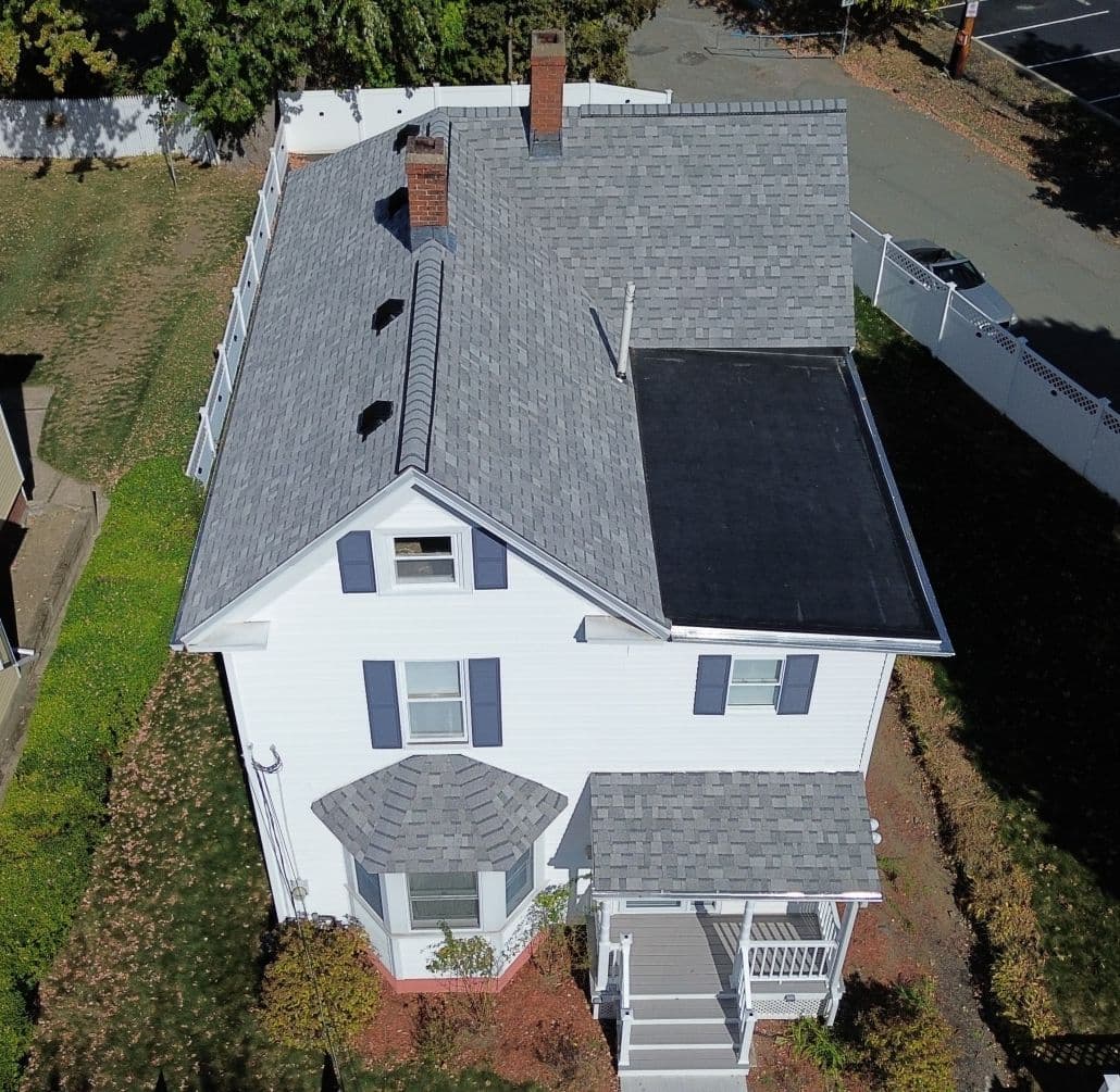 Aerial view of a two-story white house with a gray shingle roof and a small porch. The yard is partially covered with grass and bordered by a white fence.