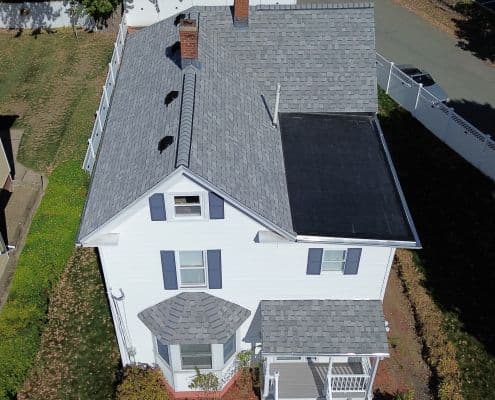 Aerial view of a white two-story house with gray shingles, blue shutters, and a small porch. A well-maintained lawn surrounds the house, creating a serene suburban setting.