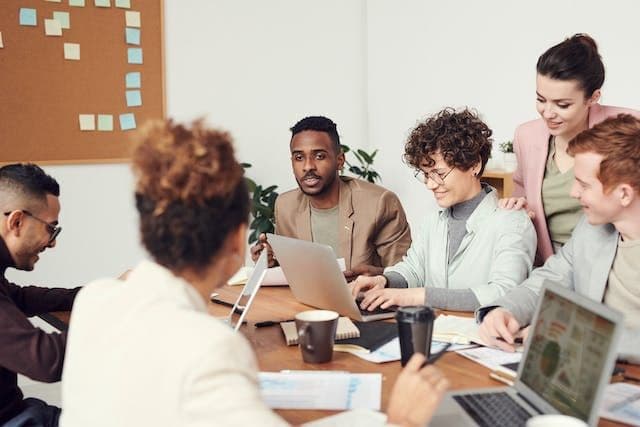 A diverse group of six people in a collaborative meeting around a table, with laptops and documents. The atmosphere is focused and engaged.