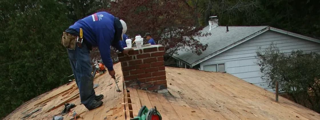 Worker repairing a roof near a brick chimney, surrounded by trees. He wears a blue shirt and tool belt, focusing diligently on the task.
