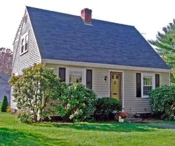 A cozy, light-brown, Cape Cod-style house with a dark roof, white windows, and a small garden. Surrounded by lush greenery on a sunny day.