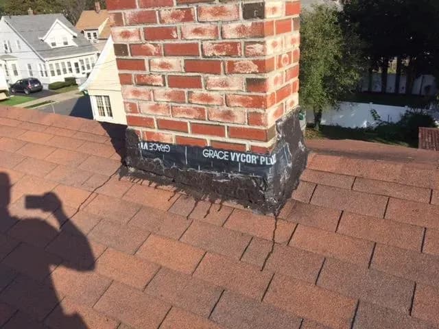 A brick chimney stands on a shingled roof with black sealant applied unevenly at its base. The words "GRACE VYCOR" are visible on the sealant. A house and trees are in the background.