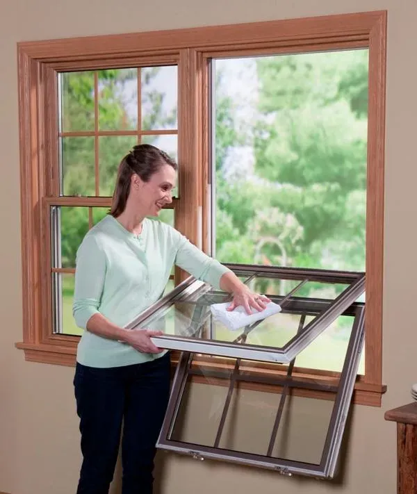 Woman in light green sweater smiles while cleaning a tilt-in window with a cloth. Background shows greenery through another window, conveying a fresh, tidy atmosphere.