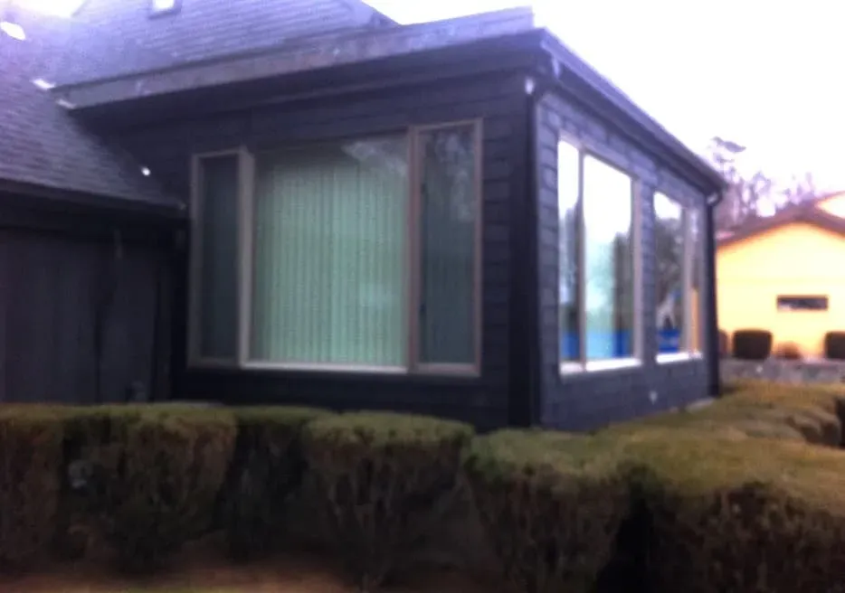 A dark-painted house extension with large windows and vertical blinds, surrounded by neatly trimmed hedges, sits under an overcast sky.