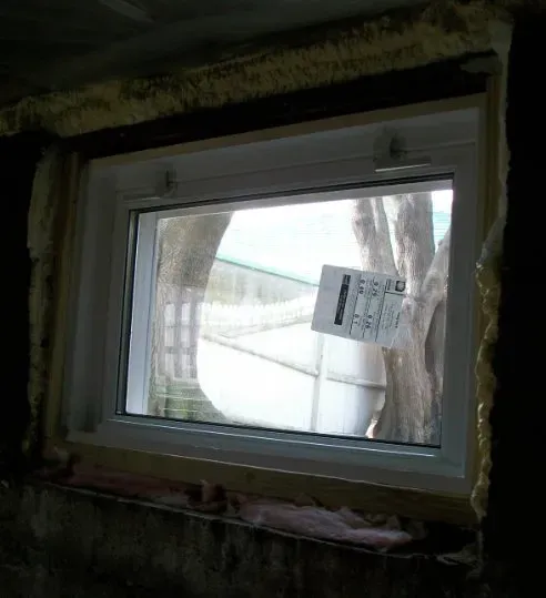 A newly installed rectangular window in a dark room, surrounded by insulation foam. A tree and fence are visible outside. A sticker is on the glass.