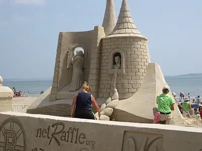 A detailed sandcastle with towers and sculptures stands on a beach. People admire the intricate designs under a clear sky, creating a lively atmosphere.