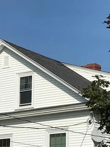 White house with a dark gabled roof and a chimney under a clear blue sky. Partial view of tree branches in the foreground, adding a natural touch.