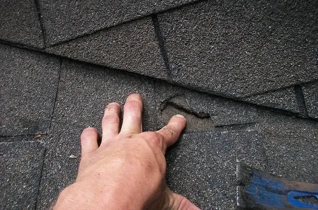 A hand inspects damaged asphalt shingles on a roof, with visible wear and a small hole. The scene conveys concern over possible roof repair needs.