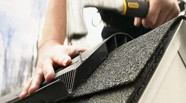 Close-up of a person using a hammer to secure a vent onto a shingled roof. The image conveys precision and focus in roofing work.