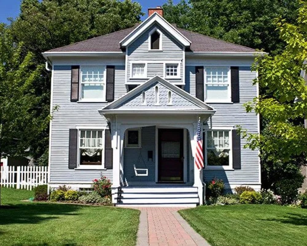Two-story gray house with white trim, dark shutters, and a small porch with a swing. A brick walkway, lush lawn, and American flag create a welcoming atmosphere.