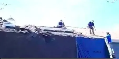 Rubble-strewn roof with three people in hard hats working, suggesting a rescue or repair effort under a bright blue sky, conveying urgency.